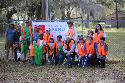 Volunteers, including children in orange safety vests holding litter grabbers and green trash bags, pose for a group photo in front of a chain-link fence with a banner reading 'Great American Cleanup' during a Keep Edisto Beautiful community cleanup event.